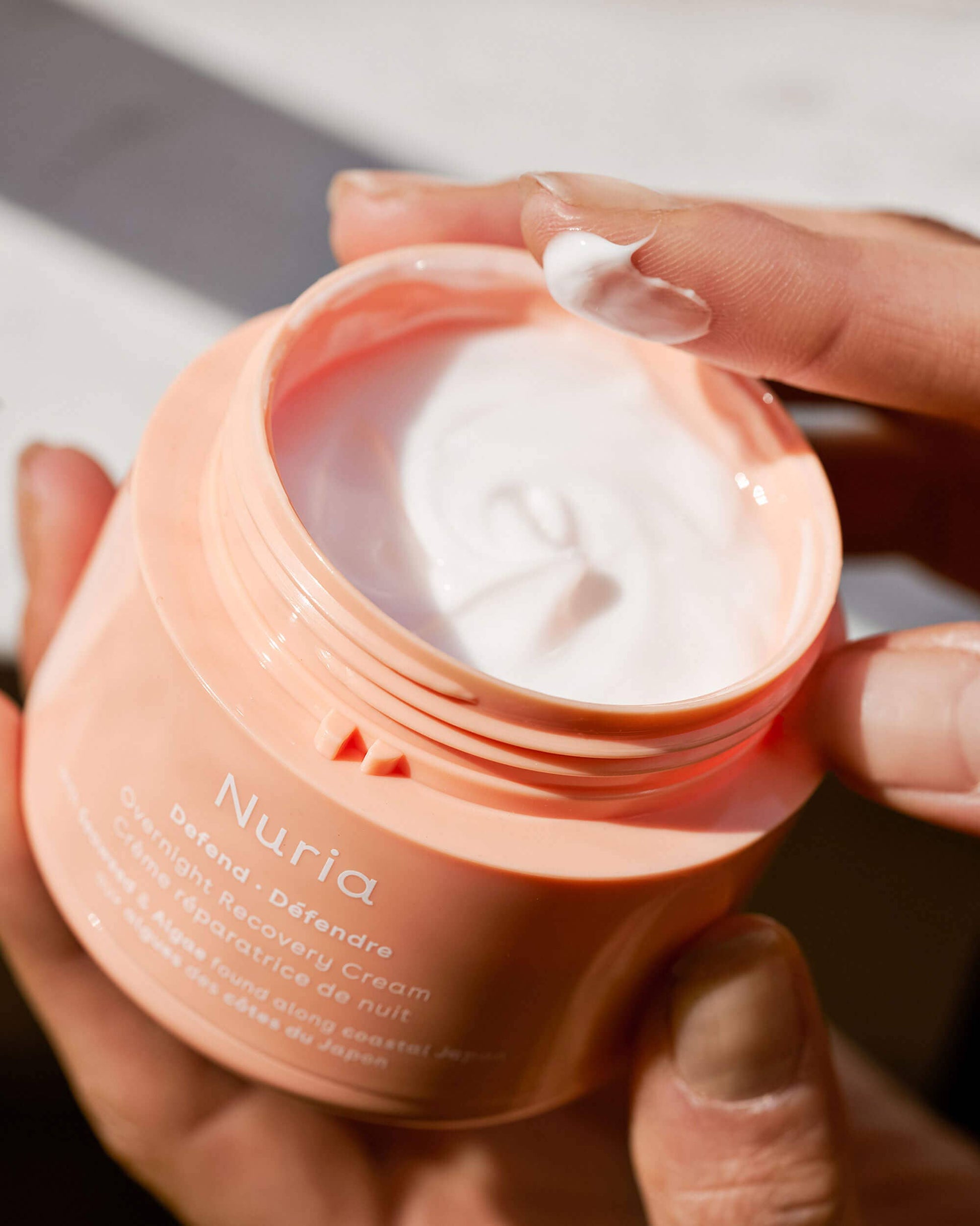 Jar of Nuria Defend Overnight Recovery Cream with a woman's hand dipped into it to show texture of the facial cream. Sold at Juniper Skincare in Edina, Minnesota.